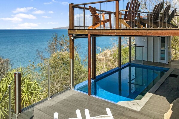 Un espace moderne aménagé au bord de la piscine, offrant une vue dégagée sur la mer, avec une terrasse en bois et un niveau supérieur vitré comprenant un coin salon.
