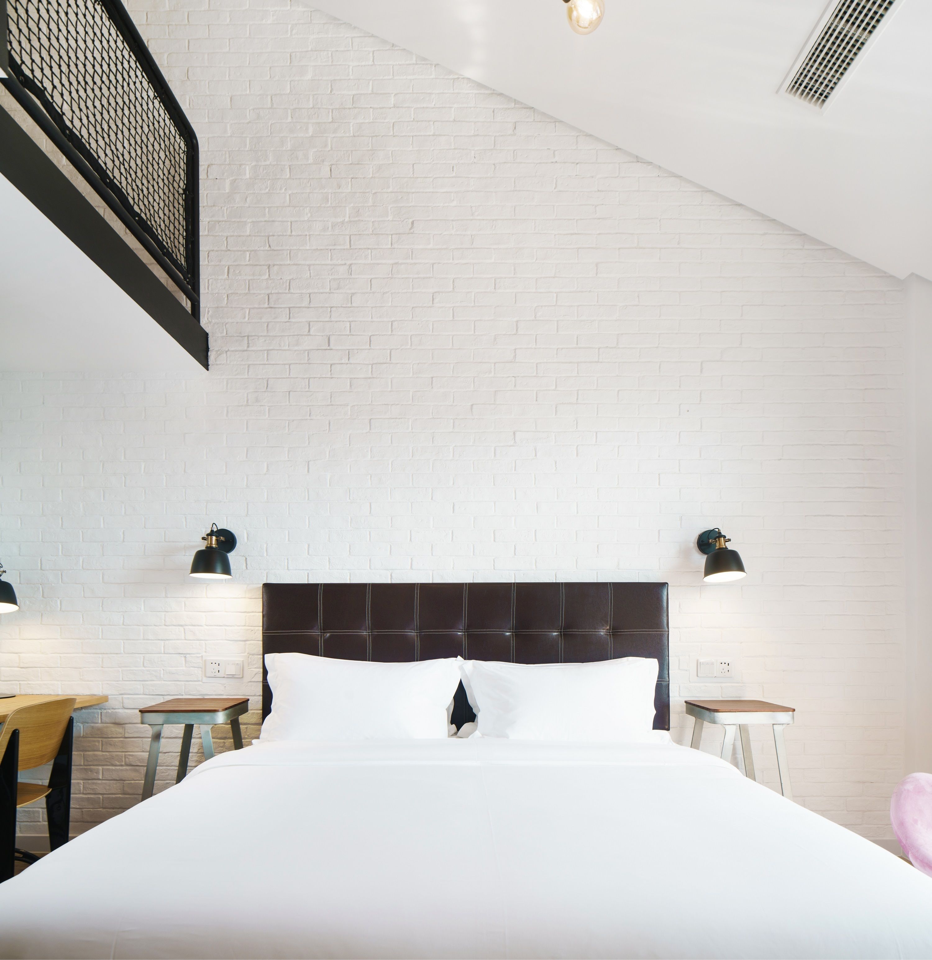 A modern hotel room with a white brick wall, a large bed with a dark headboard, two bedside tables with lamps, and a wooden chair.