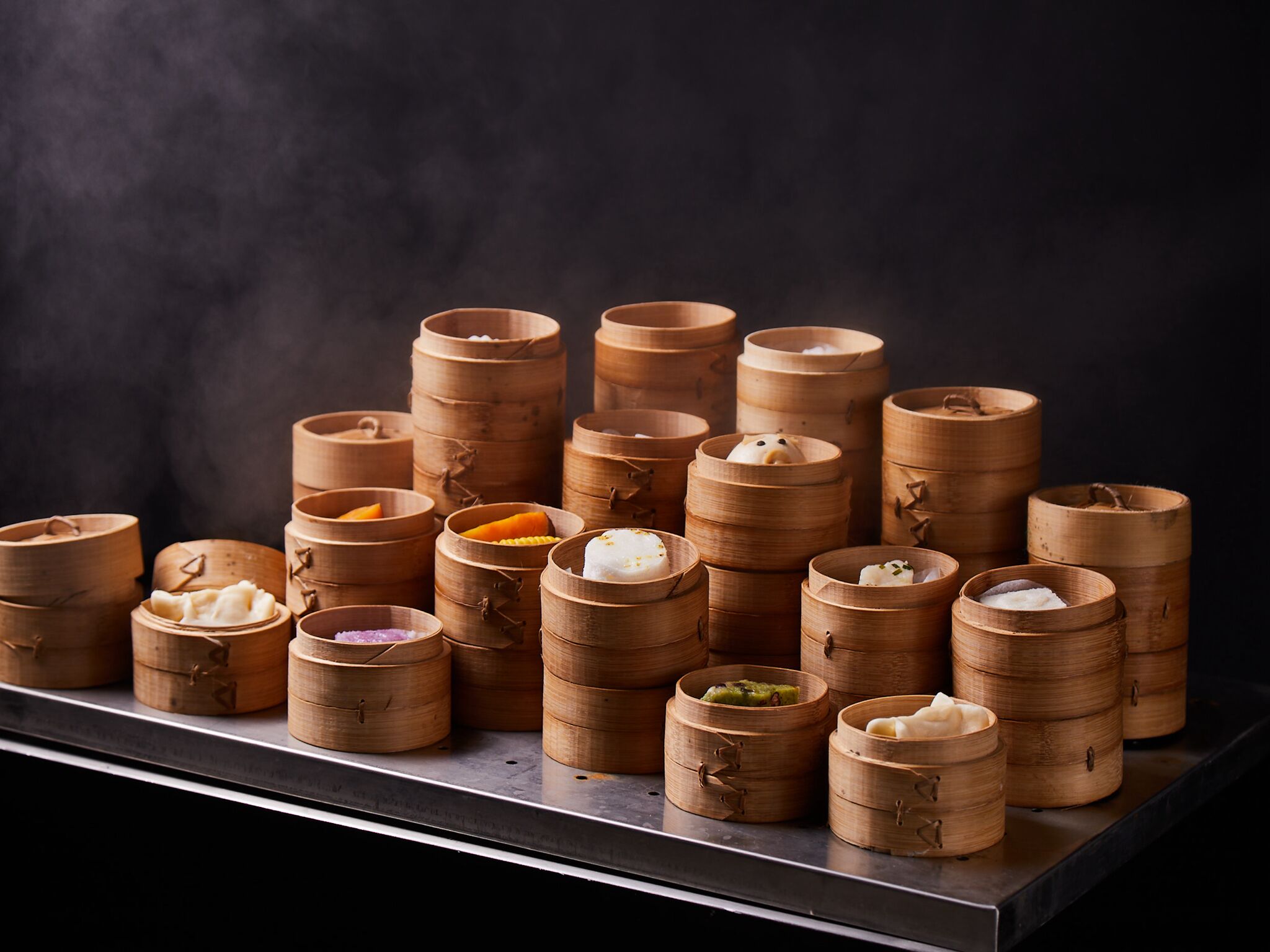 A variety of bamboo steamers with different food items, arranged on a metal shelf against a dark background.