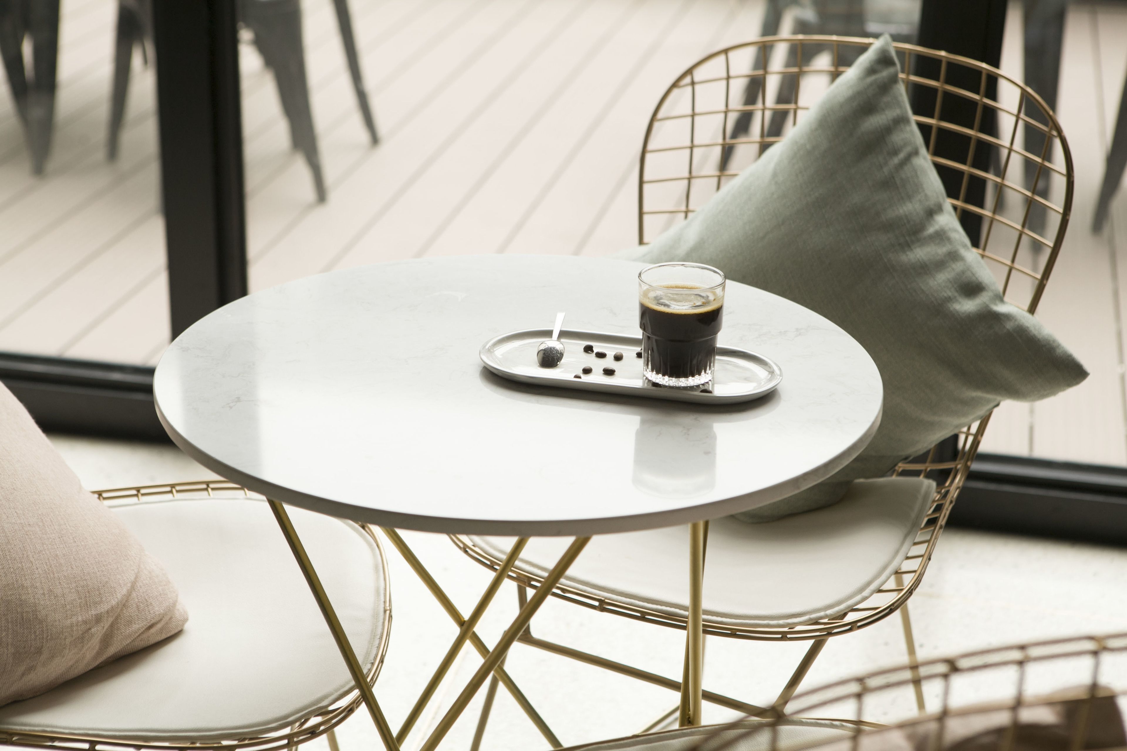 A round table with a white surface and a glass of coffee, accompanied by a tray with coffee beans and a spoon. The table is surrounded by wireframe chairs with cushions.