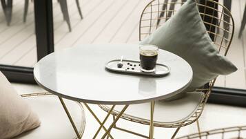 A round table with a white surface and a glass of coffee, accompanied by a tray with coffee beans and a spoon. The table is surrounded by wireframe chairs with cushions.
