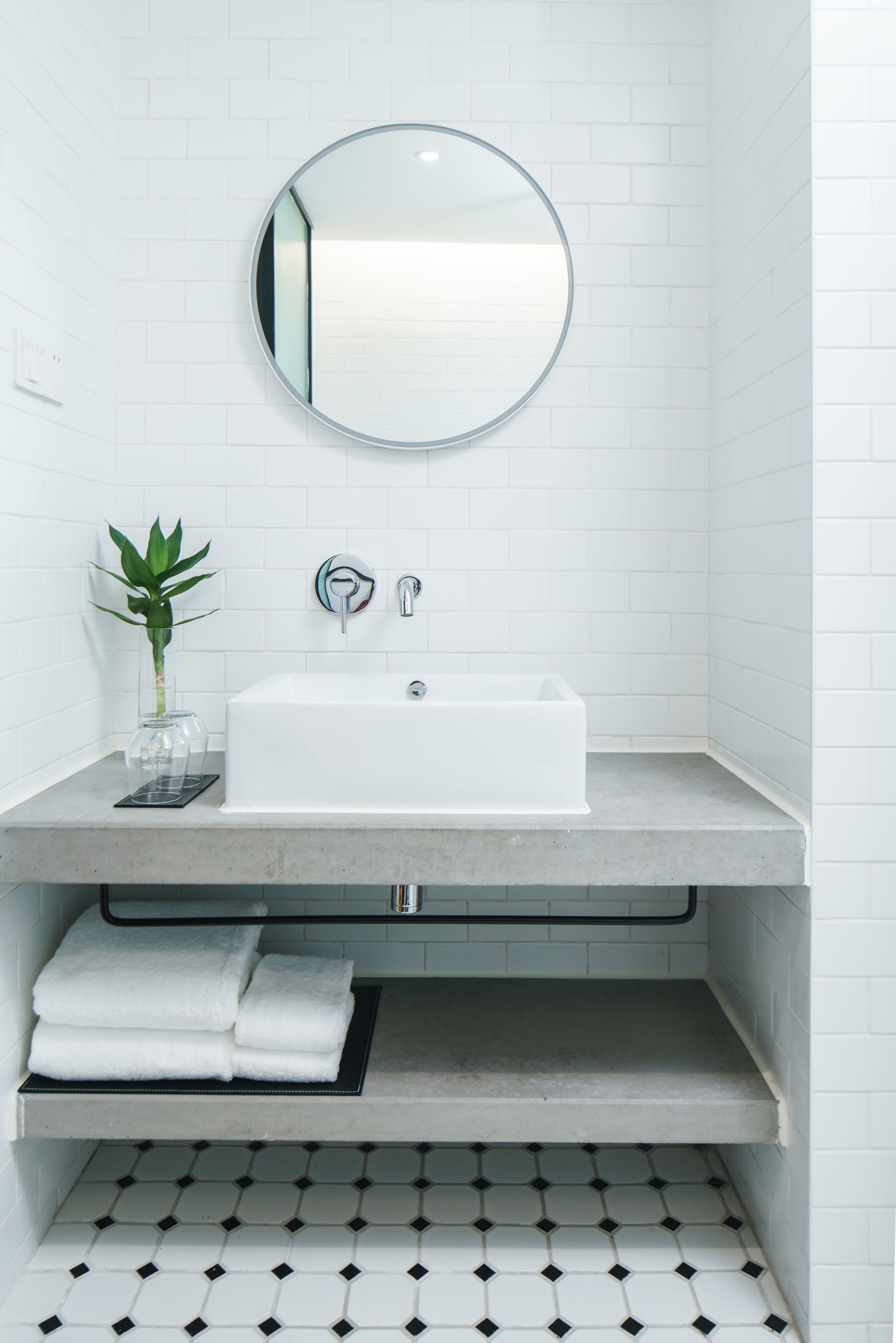 A modern bathroom with a white sink, a round mirror, a plant, and a tiled floor.