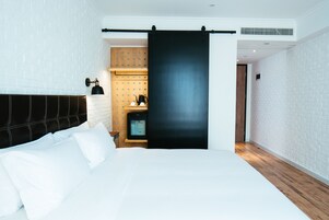 A modern hotel room with a large bed, a wooden headboard, a black sliding door, and a small kitchenette area.
