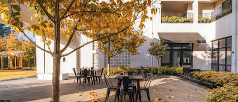 An outdoor seating area with tables and chairs, a tree with yellow leaves, and a building with balconies and windows.