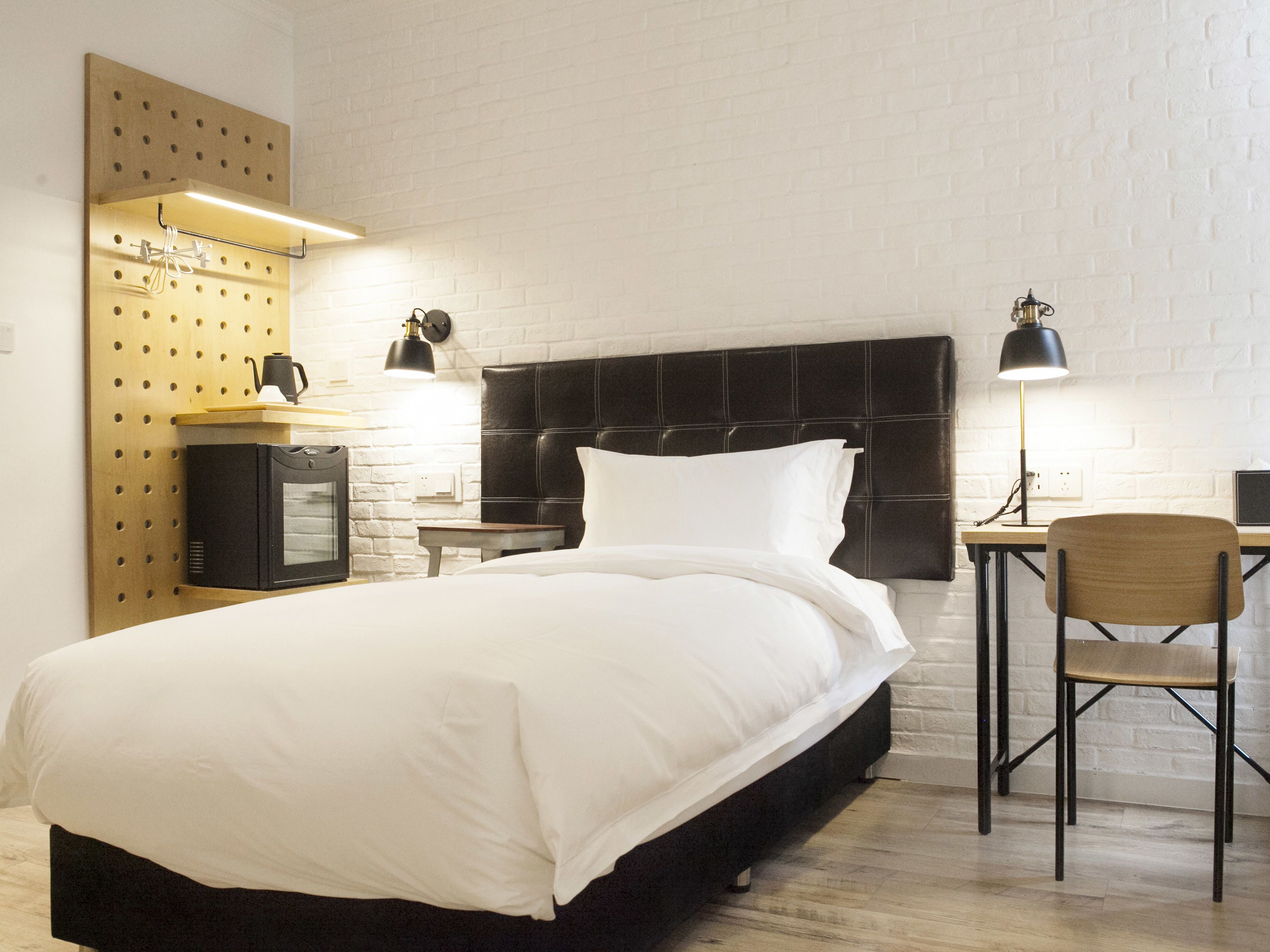 Een netjes opgemaakt bed met een zwart hoofdeinde, een klein bureau met een stoel, en een wandplank met een waterkoker en een koffiepot.