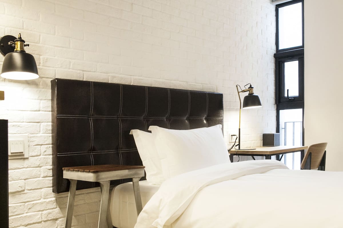 Habitación de hotel con pared de ladrillo blanco, cabecero negro, mesita de noche con lámpara y una silla.