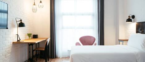 A hotel room with a wooden desk, two chairs, a bed, and a large window with curtains.