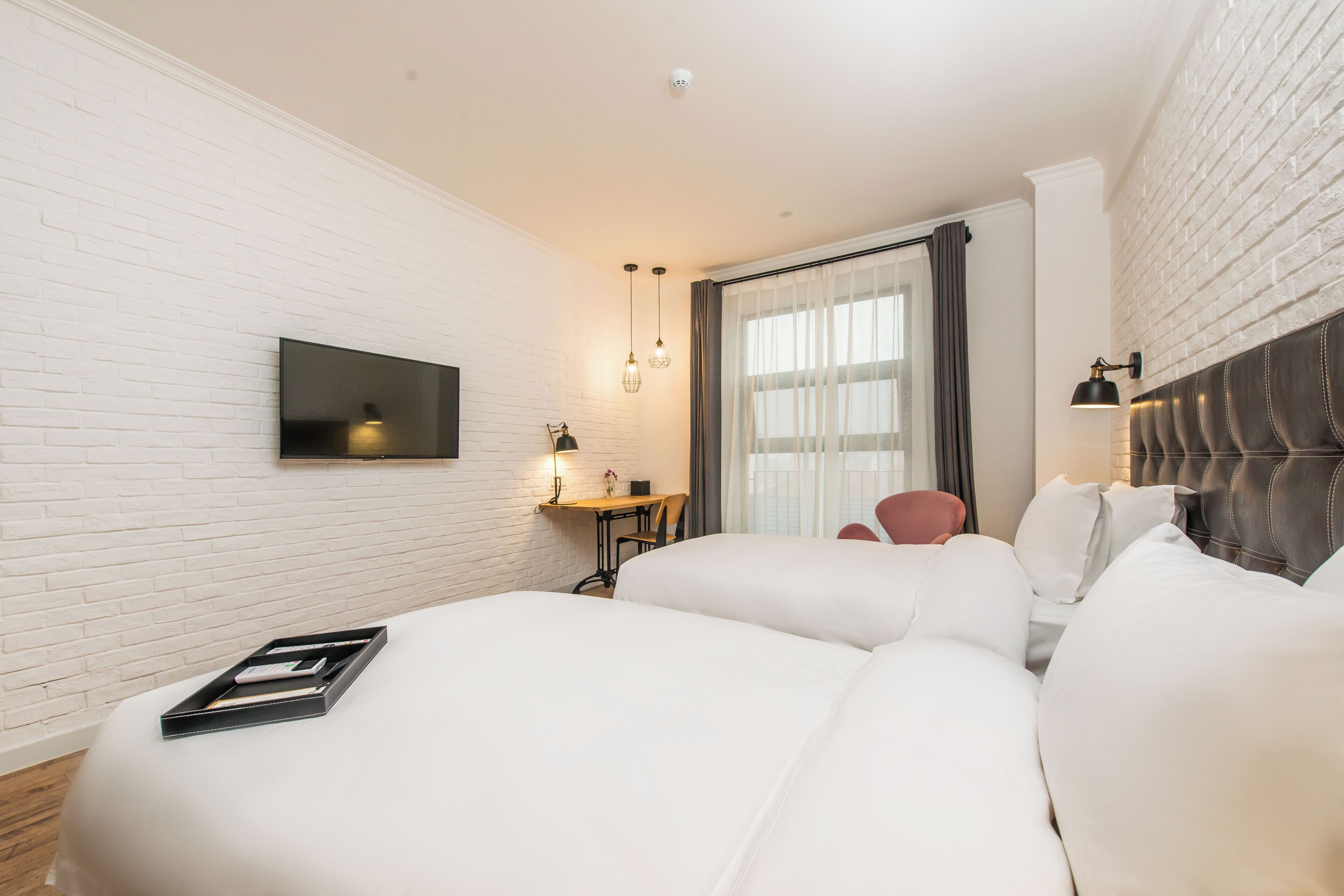 A hotel room with a white brick wall, a flat-screen TV mounted above a desk, a bed with white bedding, a red chair, and a window with curtains.