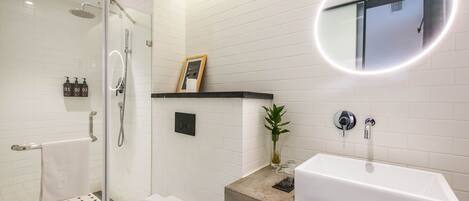 Un baño moderno con ducha de vidrio, inodoro blanco, lavabo rectangular y espejo con marco circular.