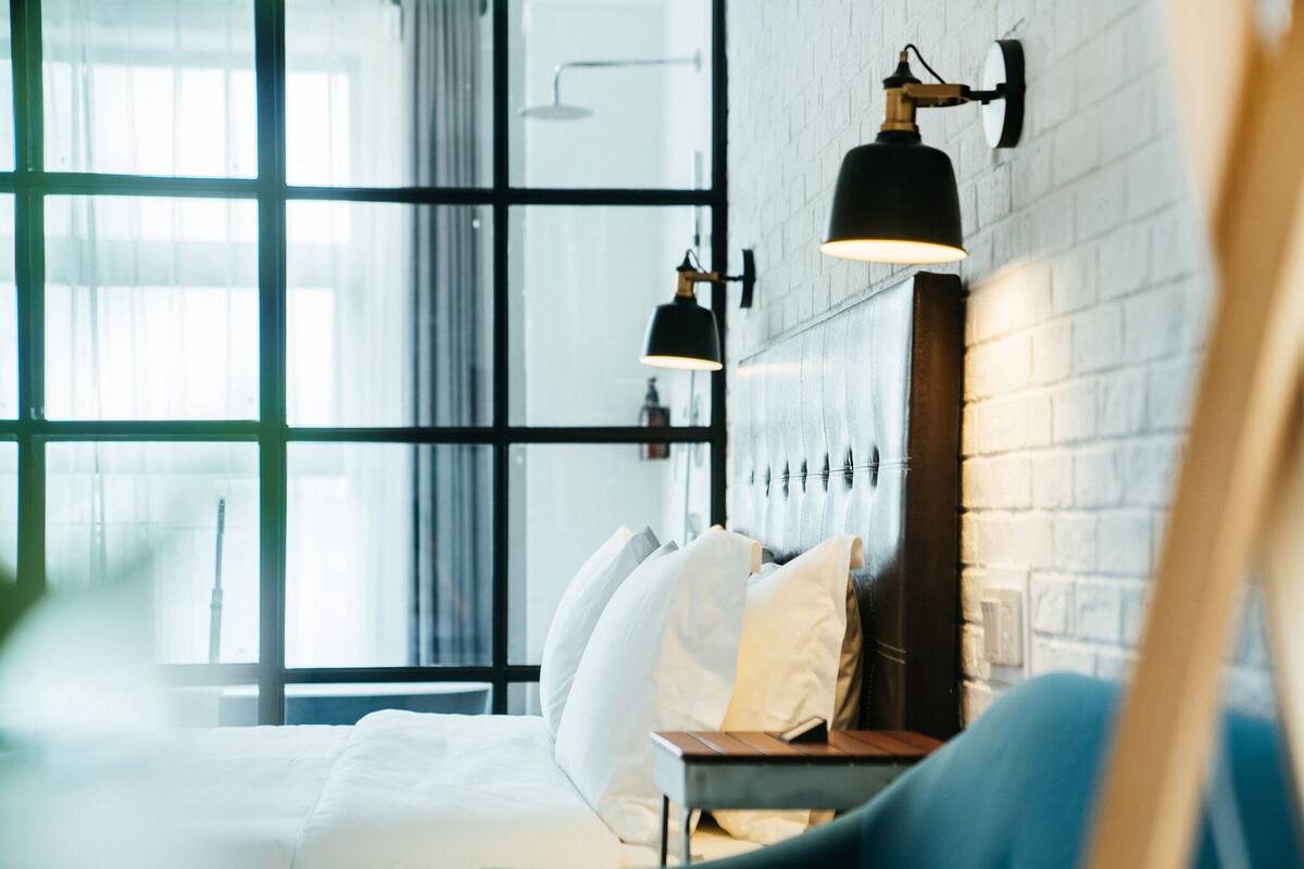 Habitación de hotel moderna con un gran ventanal, una cama con s ábanas blancas, un sillón azul y dos lámparas fijadas a la pared.