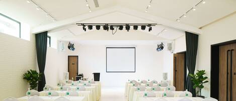A conference room with rows of tables and chairs, a projector screen, and ceiling-mounted lighting.
