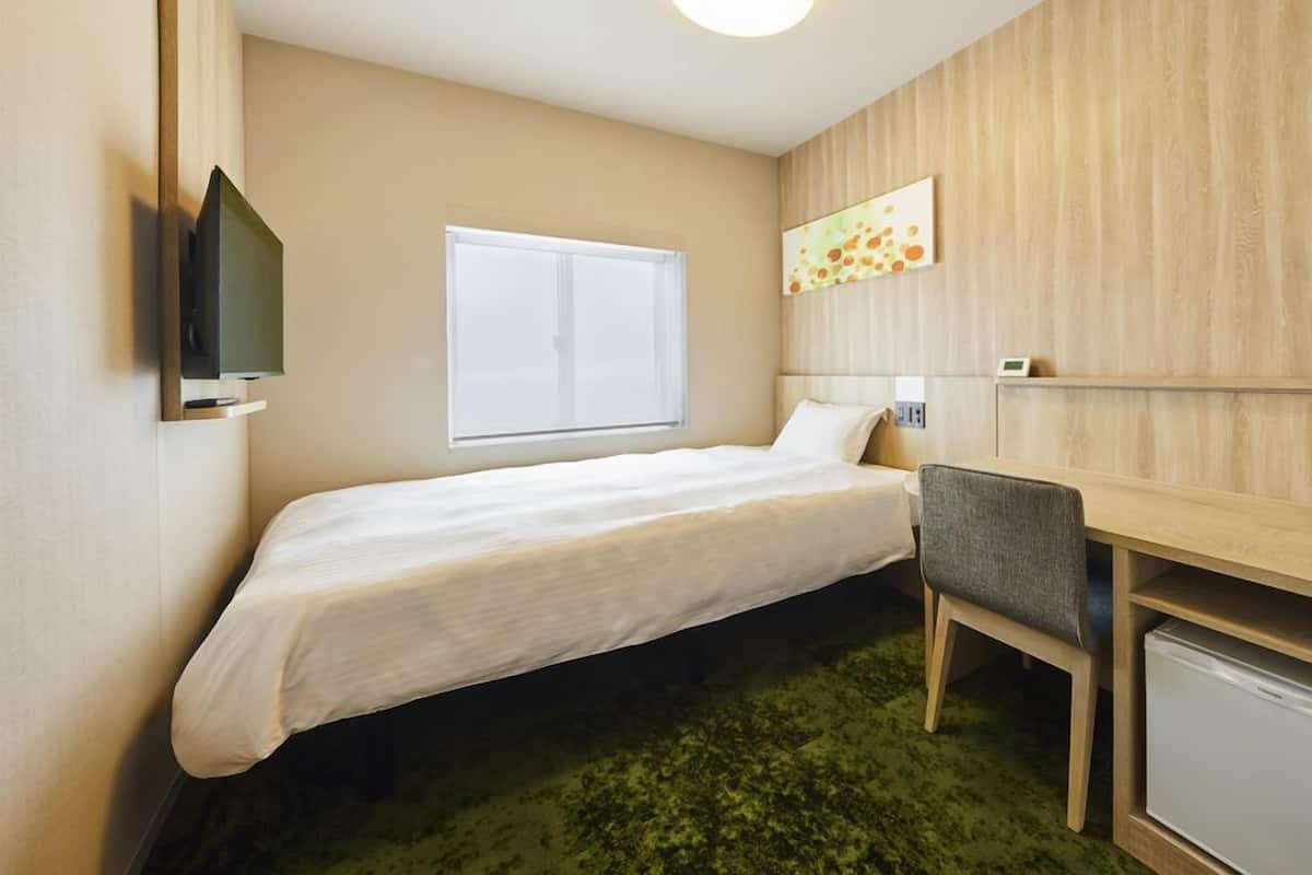 A single bed with a white mattress, a small desk with a chair, a television mounted on the wall, and a window with closed blinds.