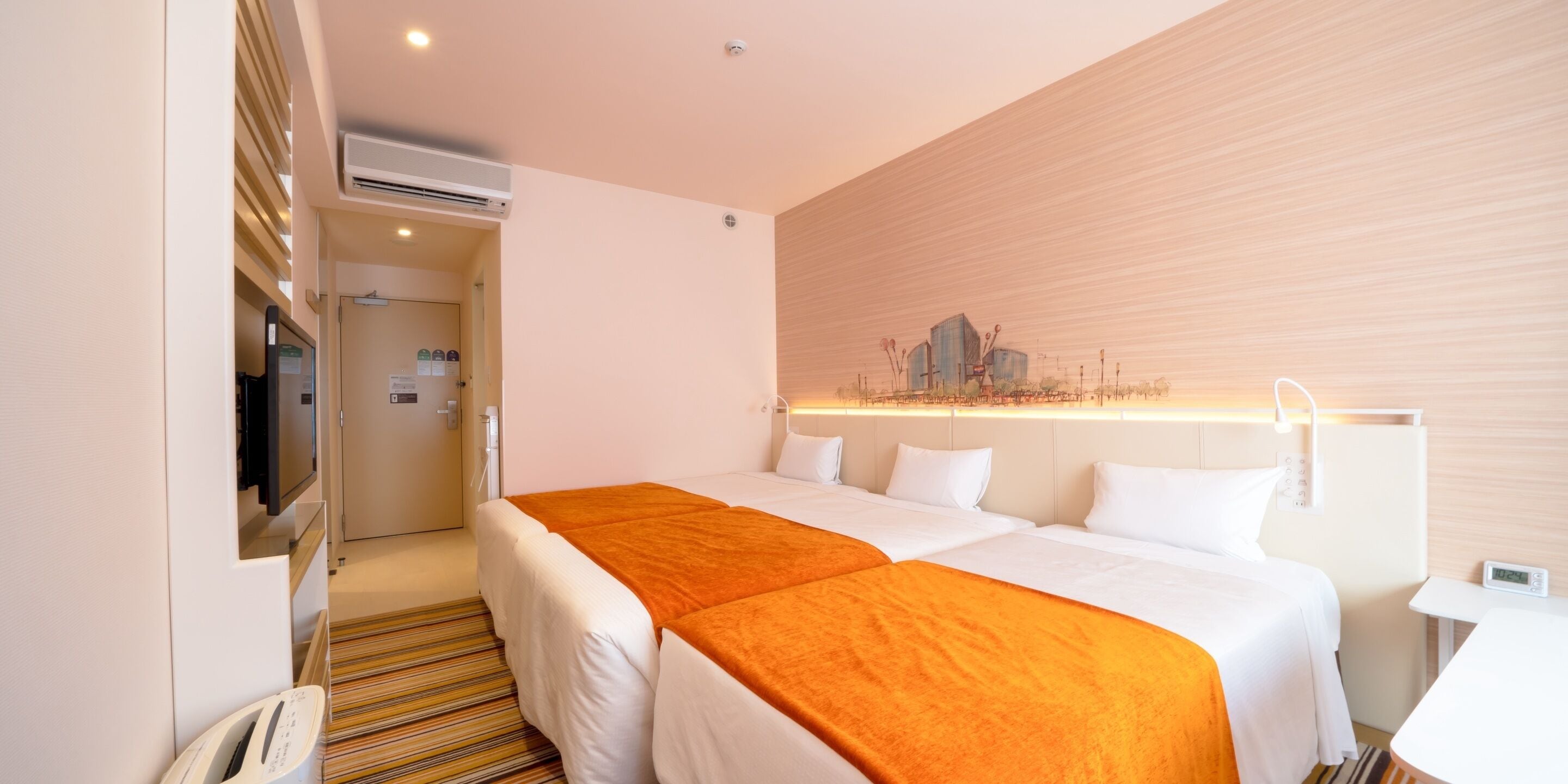 Habitación de hotel con dos camas, un televisor, un equipo de aire acondicionado y una obra de arte con un paisaje urbano en la pared.