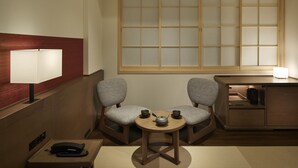 A traditional Japanese-style room with a tatami mat floor, a low wooden table with a tea set, two cushioned chairs, a sliding shoji screen, and a bedside table with a lamp. - Ours Inn Hankyu (Tokyo)