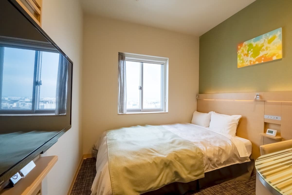 A hotel room with a bed, a bedside table with a phone, a TV, and a window with a city view.