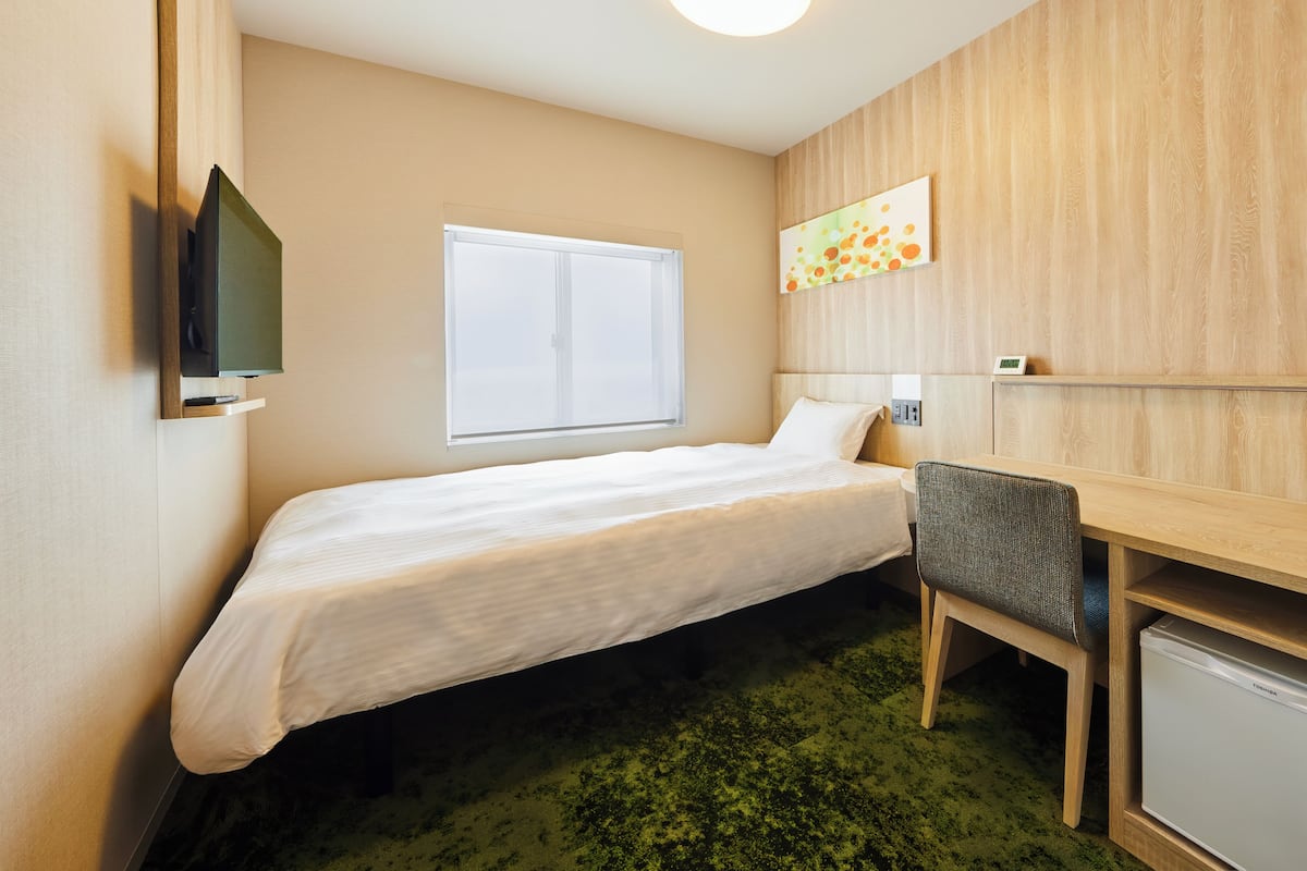 A hotel room with a bed, a desk, a chair, a television, a window, and a small refrigerator.