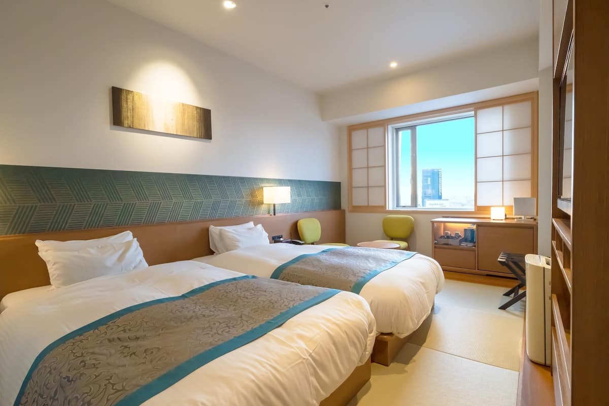 A hotel room with two beds, a desk, a chair, a window with a city view, and a bedside table with a lamp.
