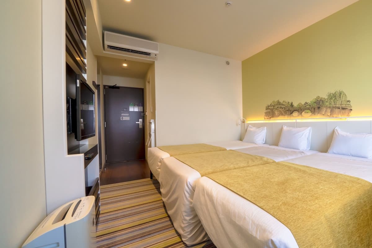 A hotel room with two beds, a small table, a TV, and a wall-mounted air conditioner.