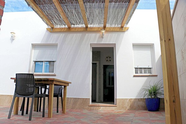 Un patio con mesa y sillas de madera, una planta en maceta y una zona de estar cubierta con techo estampado.