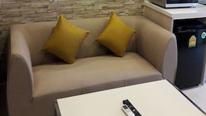Ett modernt vardagsrum med en vägg som har en stenliknande struktur, en beige soffa med gula kuddar, ett vitt soffbord med en fjärrkontroll och en mikrovågsugn placerad på en svart bänkskiva.