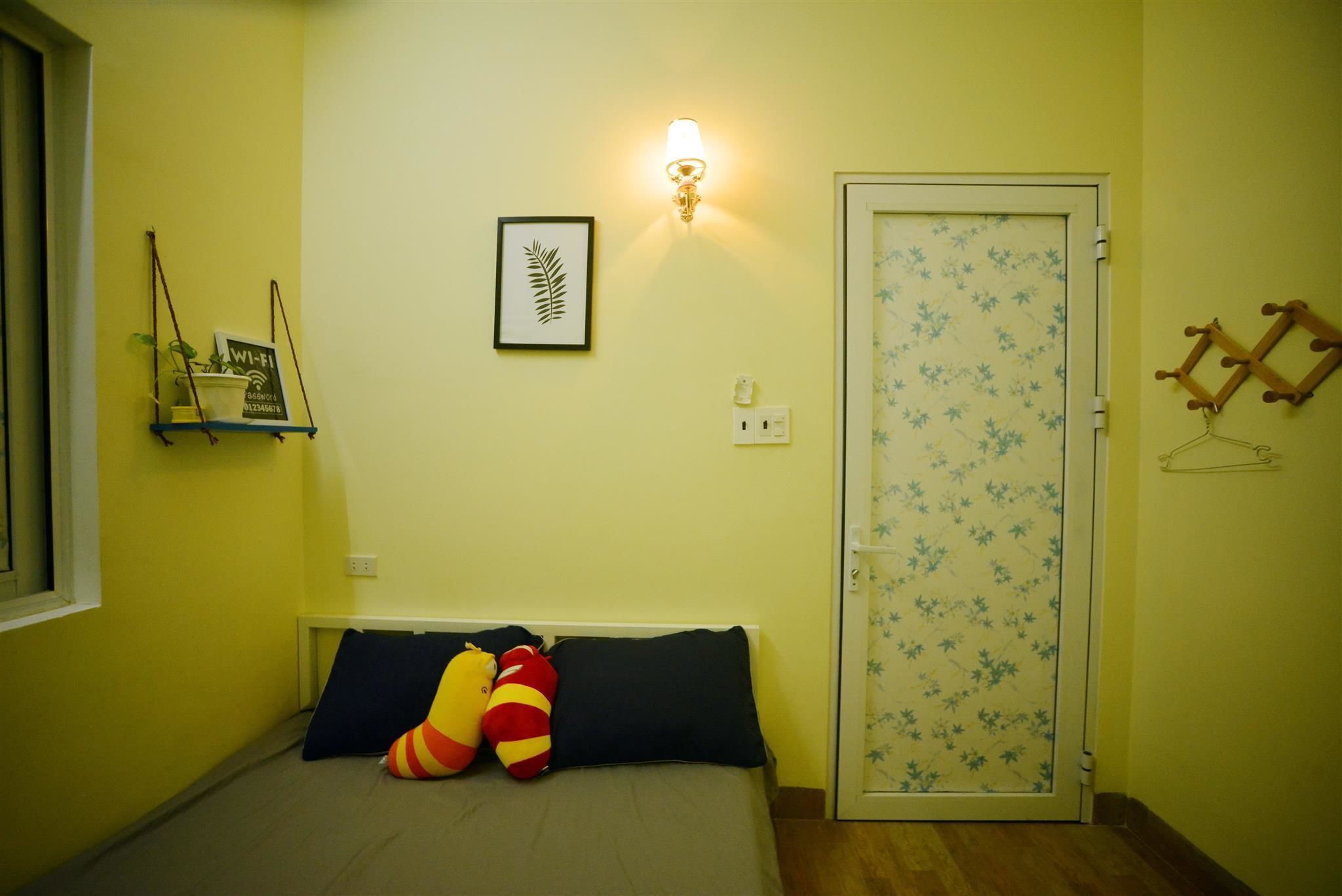Quarto com cama, almofadas, luminária na parede, porta com padrão floral e uma prateleira pequena com um sinal de Wi-Fi.