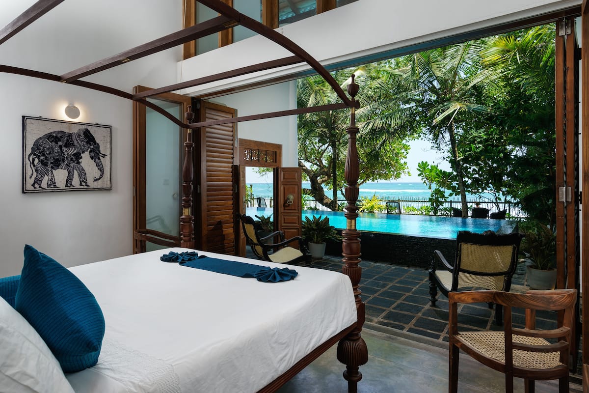 A bedroom with a large bed, a view of a pool and palm trees, and a wooden ceiling structure.
