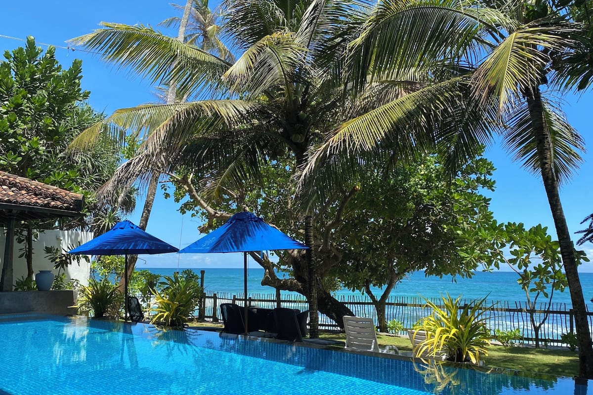 A poolside area with a view of the ocean, palm trees, and blue umbrellas.