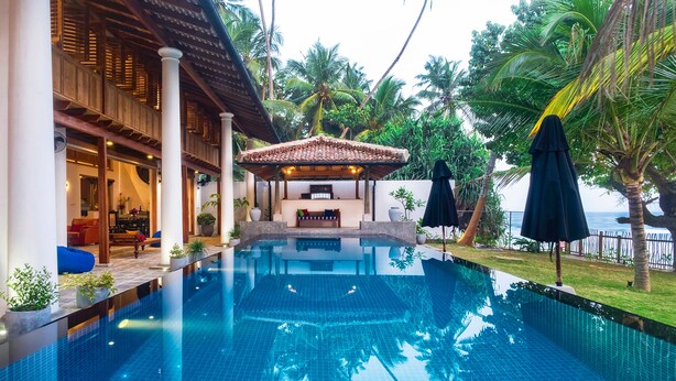 A swimming pool with a tiled deck, surrounded by palm trees and a covered seating area. - Royal Indigo Villa (Unawatuna)