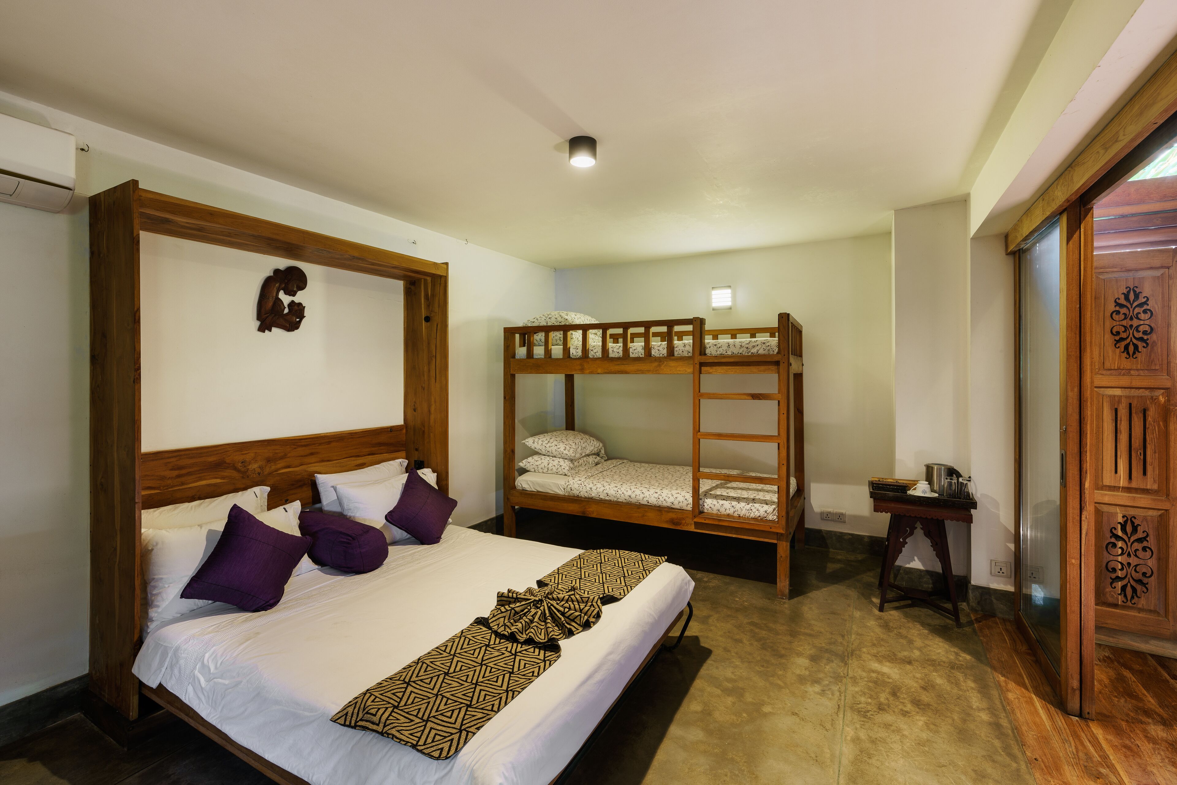 A bedroom with a large bed, a smaller bunk bed, wooden headboard, a bedside table with a lamp, and a wardrobe with a decorative panel.