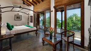 A bedroom with a large bed, a wooden table, and a chair, a balcony with a view of greenery and the sea. - Royal Indigo Villa (Unawatuna)