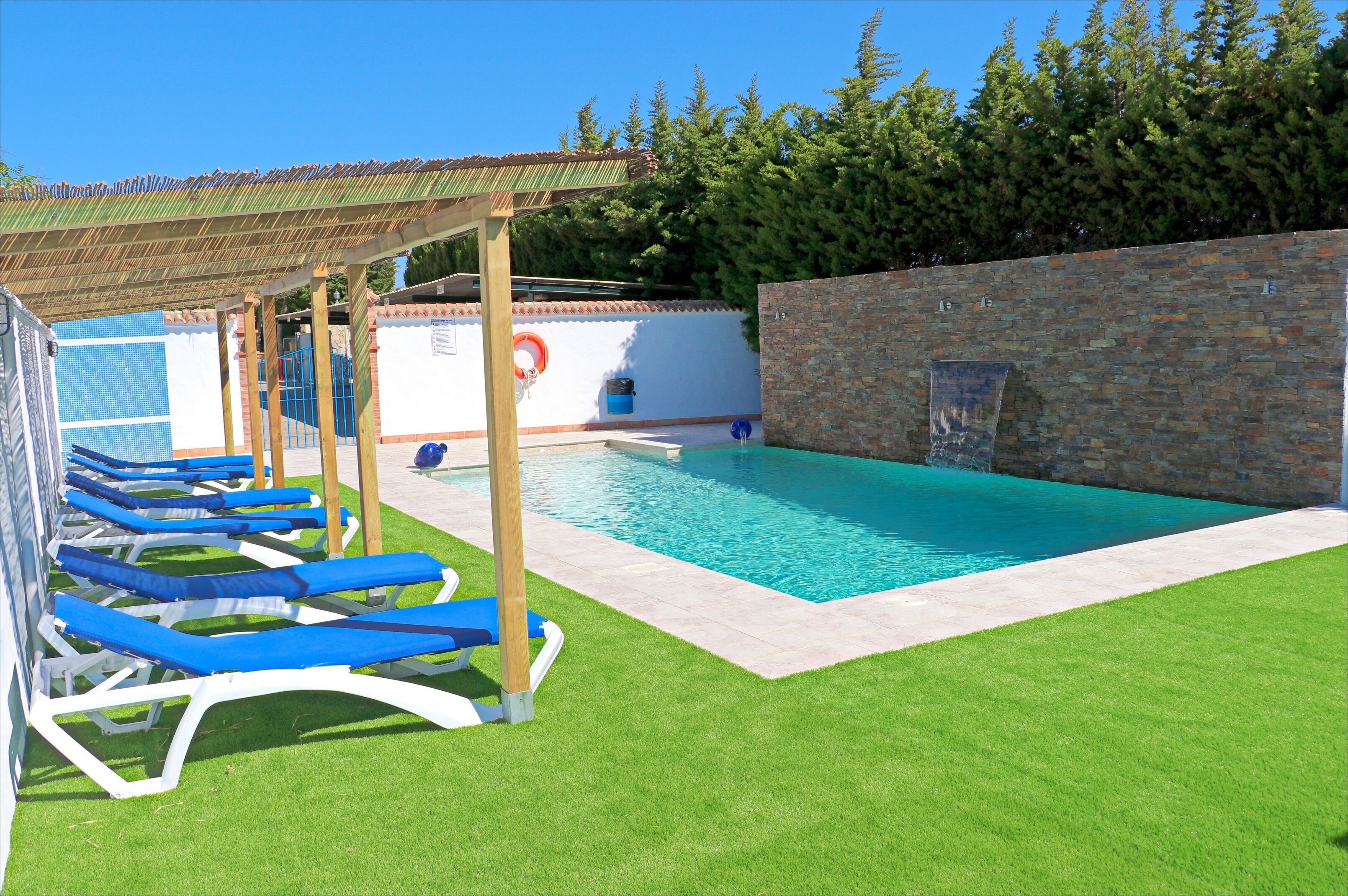 A swimming pool with blue lounge chairs, a pergola, and a stone wall.