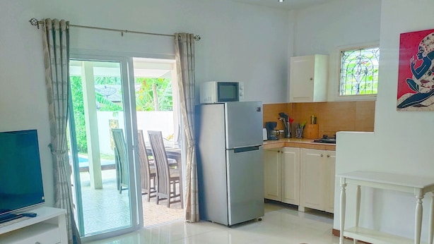 A modern living room with a glass-top coffee table, brown sofas, a flat-screen TV, and a kitchen area with a refrigerator, microwave, and cabinets. - Wirason 2-Bedroom Pool Villa (Koh Samui)