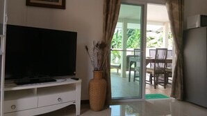 A living room with a flat-screen TV on a white stand, a large woven vase, and a sliding glass door leading to an outdoor patio with a table and chairs. - Wirason 2-Bedroom Pool Villa (Koh Samui)