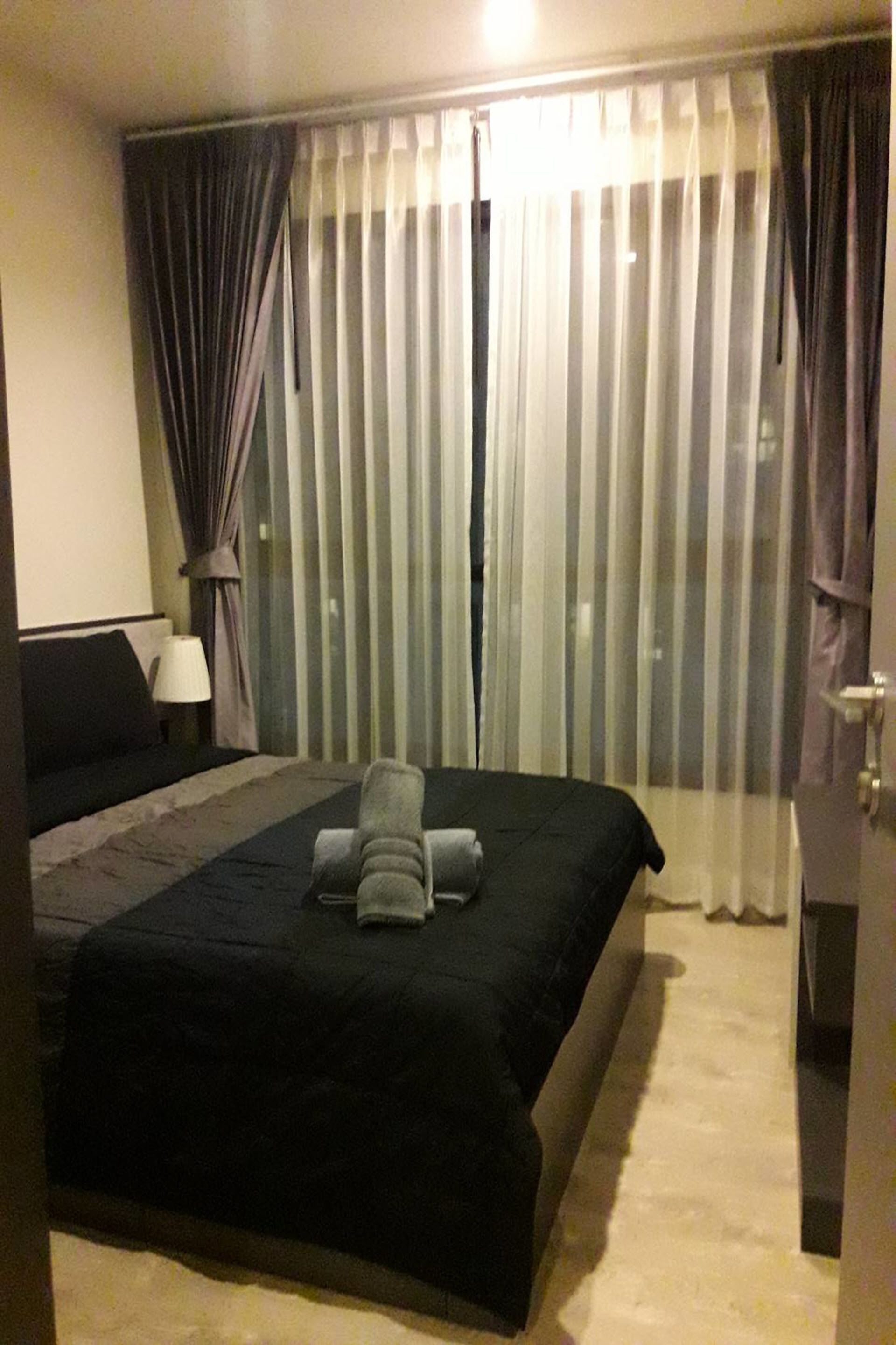 A hotel room with a large bed, a nightstand with a lamp, and a view through sheer curtains.