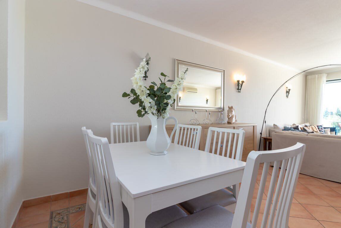 A dining area with a white table, four chairs, a vase with flowers, a mirror, and a sofa in the background.