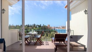 A balcony with a table and chairs, a bench, and a view of the sea and buildings.