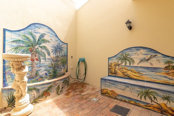 Une pièce ornée d’une fresque murale en carreaux représentant un paysage tropical, d’un piédestal blanc et d’un banc décoré d’une fresque similaire.