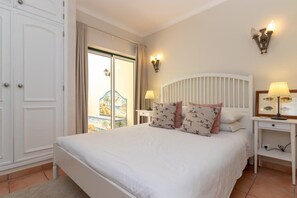 A bedroom with a bed, a nightstand, a lamp, a wardrobe, and a view of the outside.