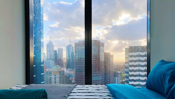 Een slaapkamer met een bed, een raam met uitzicht op de stad en een blauw kussen.