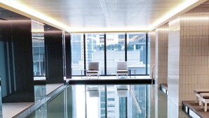 Een binnenzwembad met helder water, twee stoelen en een uitzicht op gebouwen door grote ramen.