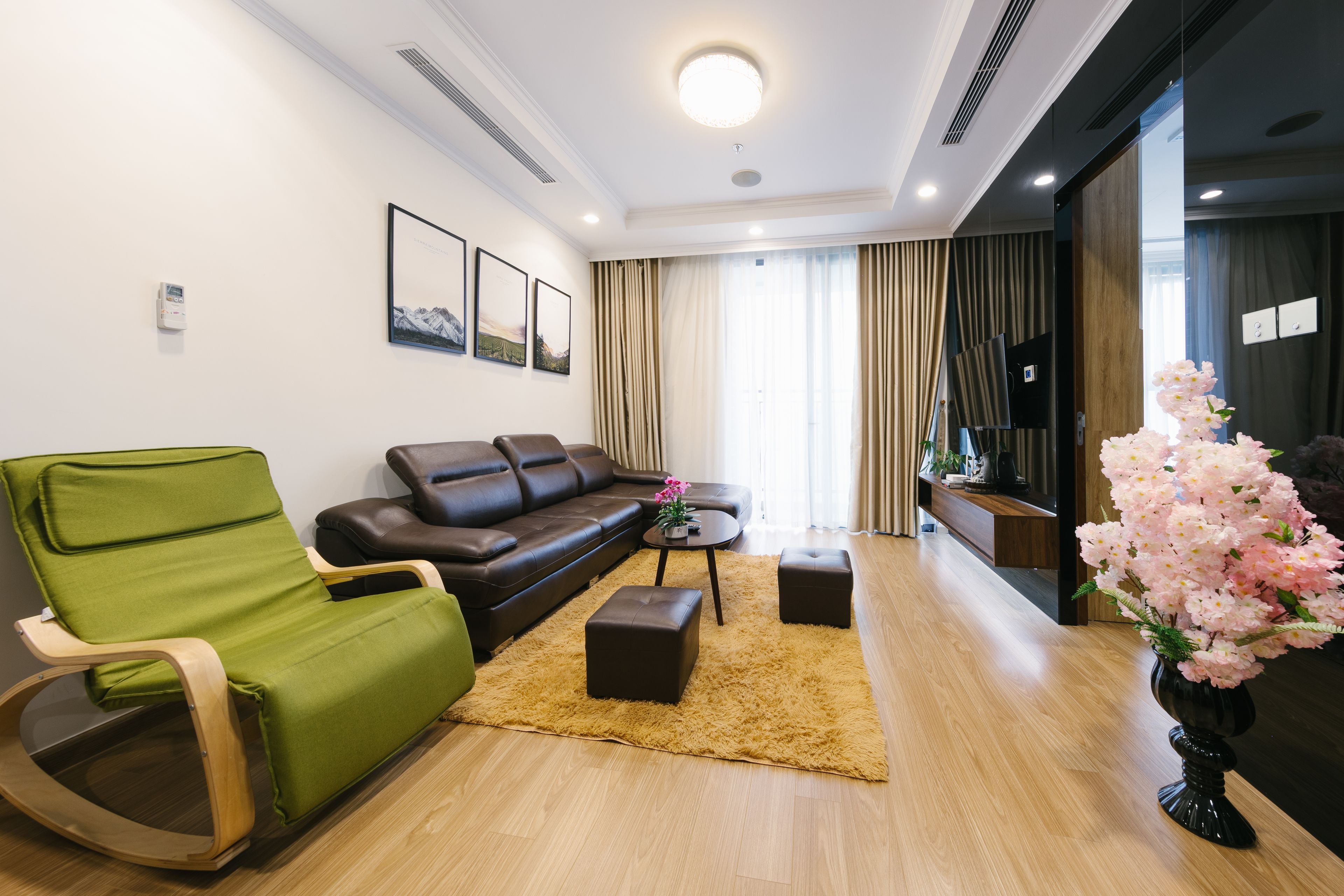 A modern living room with a green rocking chair, a brown leather sofa, a small coffee table, and a decorative vase with pink flowers.