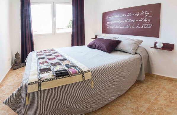 Una cama bien tendida con colcha a rayas y una colcha adicional, una obra de arte en la pared con texto, un estante pequeño con un adorno, y una estatua en el suelo.