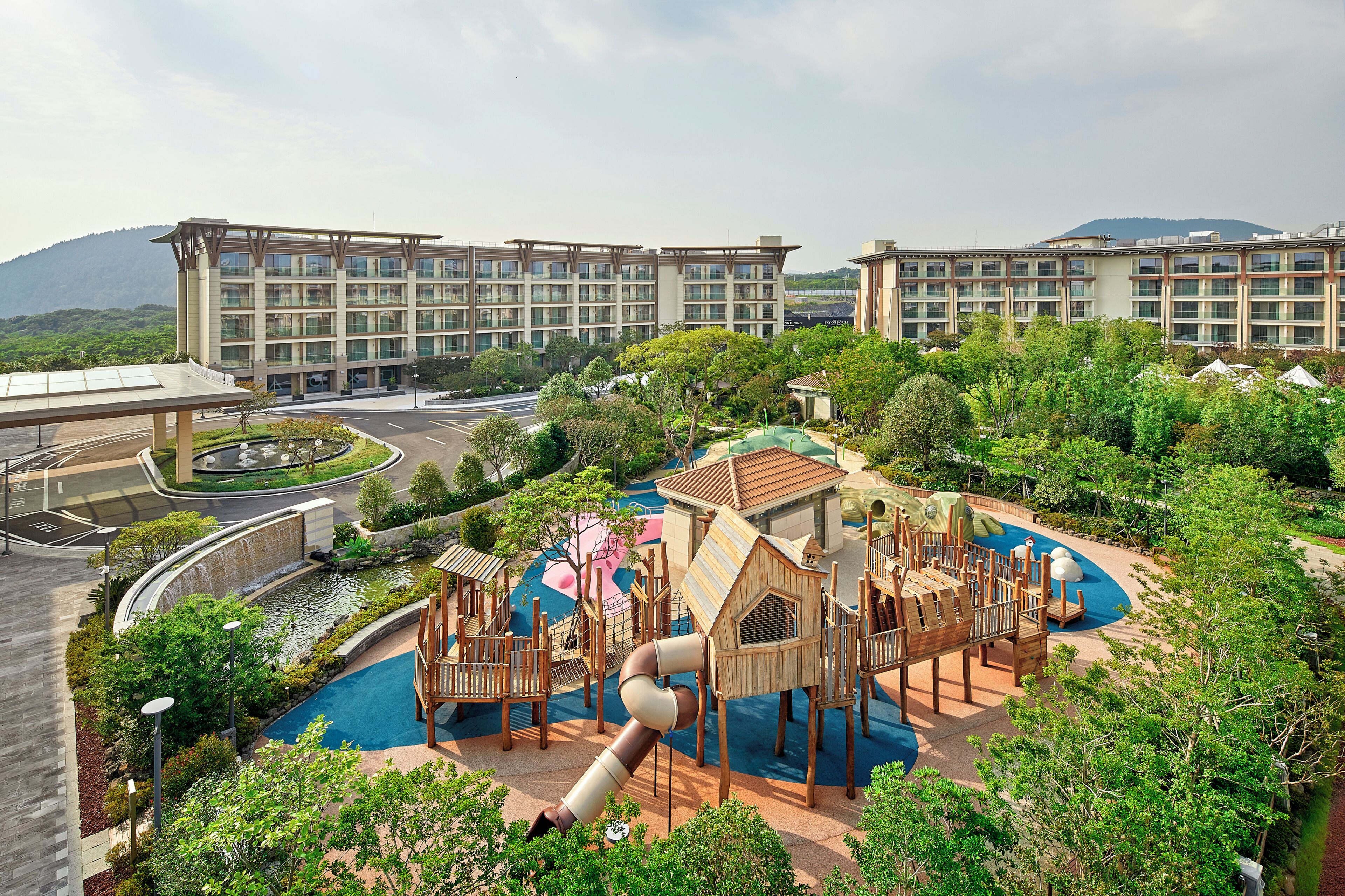 A playground with slides and climbing structures, surrounded by greenery and a pool.