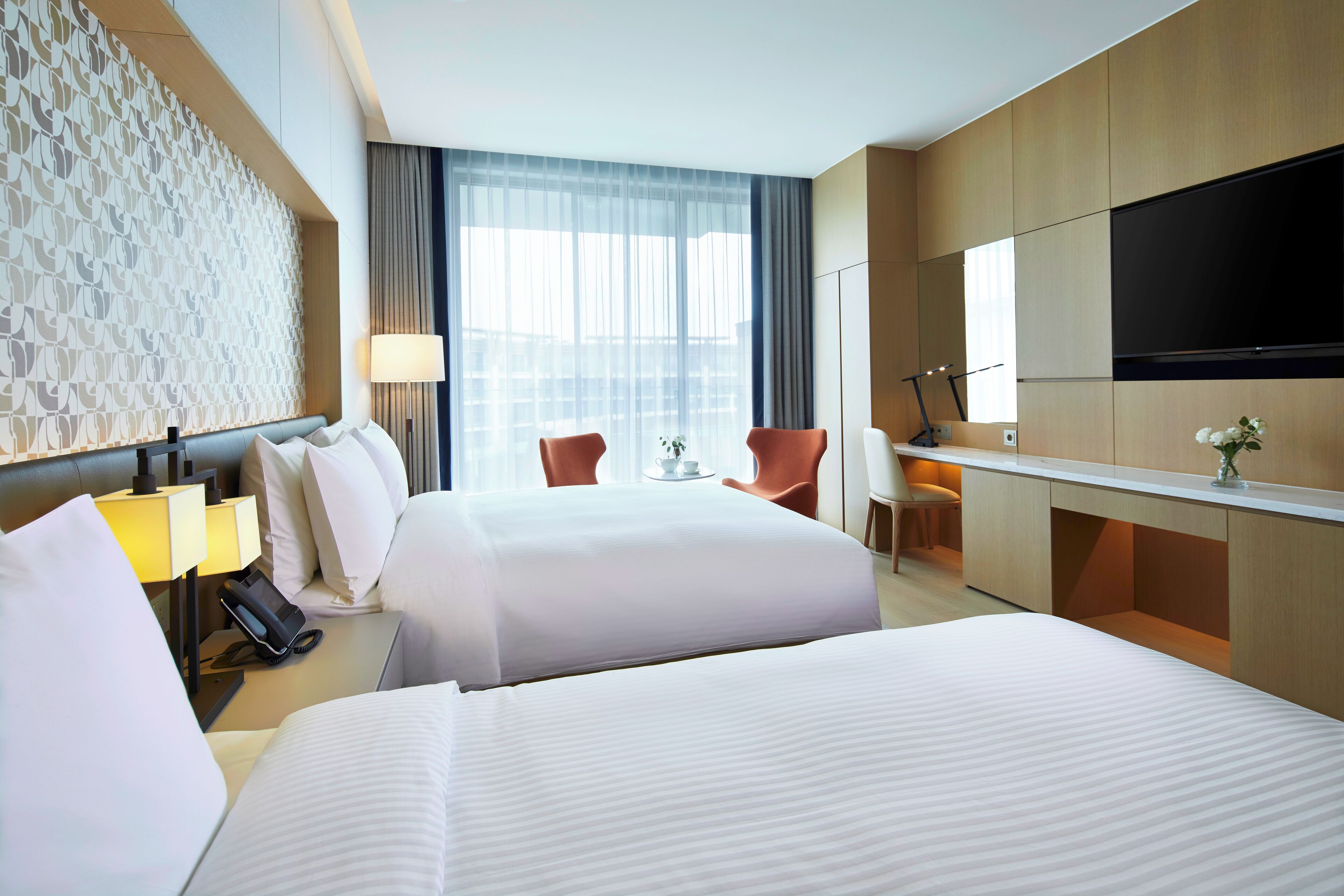 A hotel room with two beds, a desk, a chair, a TV, and a large window with curtains.