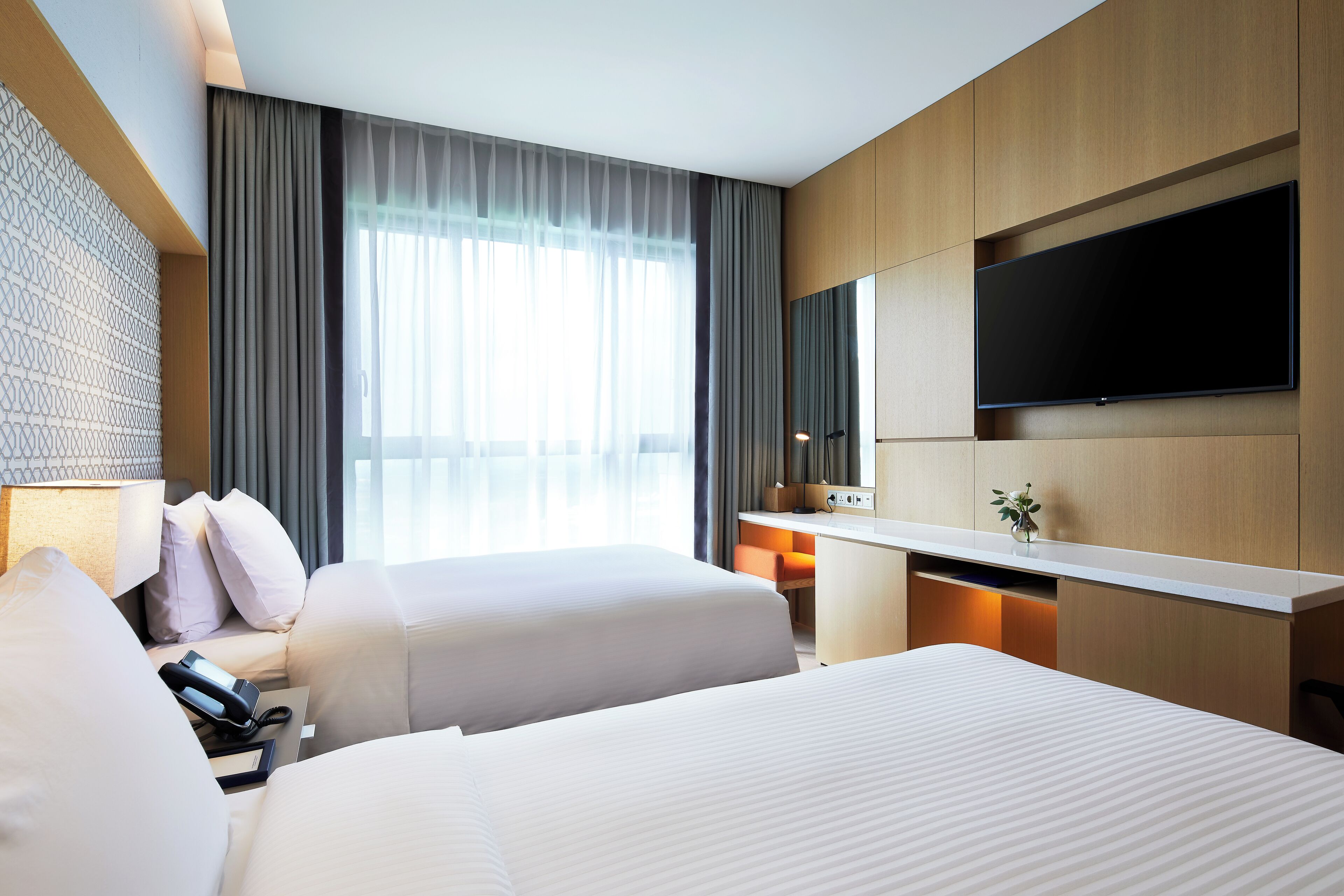 A hotel room with two beds, a desk, a flat-screen TV, and a large window with curtains.