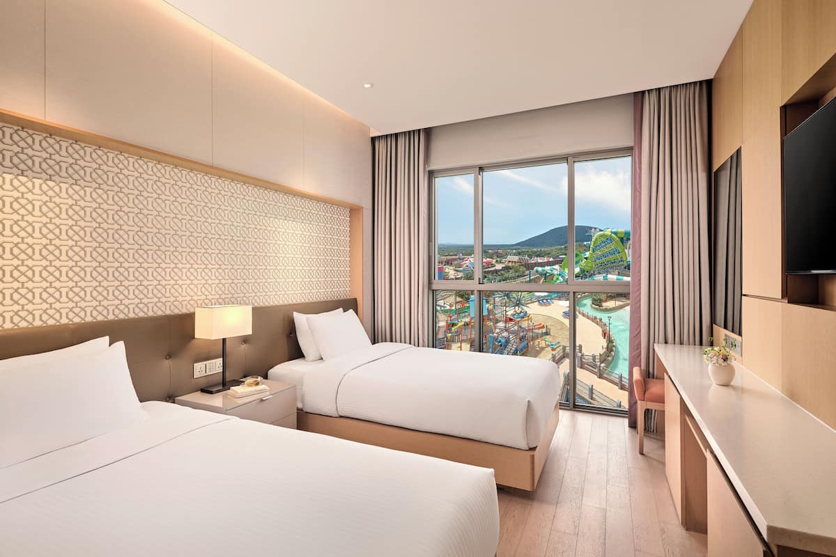 Une chambre d’hôtel avec deux lits, un bureau et une grande fenêtre donnant sur un paysage urbain et une étendue d’eau.