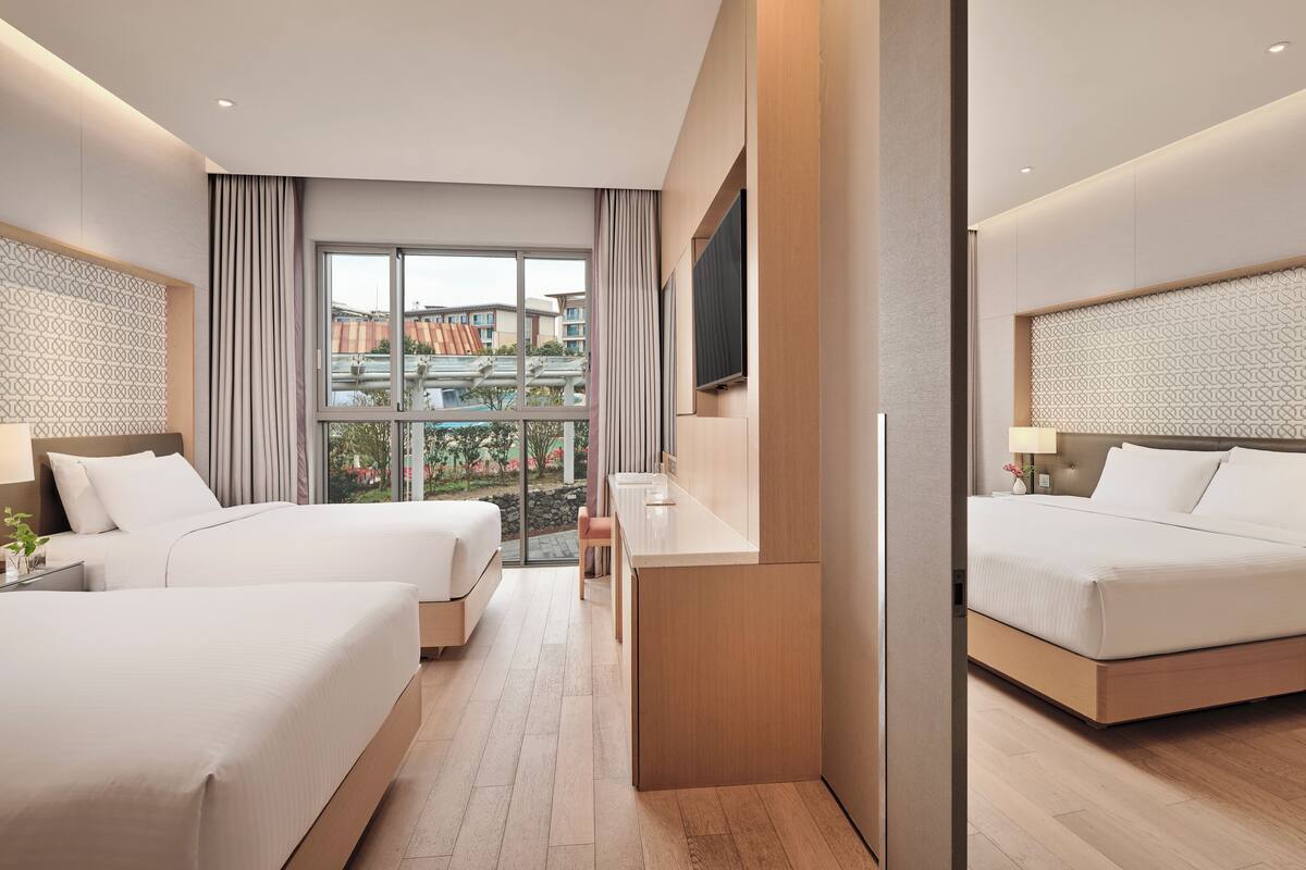 Une chambre d’hôtel moderne avec deux lits, une grande fenêtre donnant sur des immeubles et un téléviseur à écran plat fixé au mur.