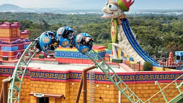 Una montaña rusa con una gran mascota de dragón, un descenso pronunciado y una vista del paisaje urbano al fondo.