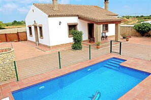 A swimming pool with a stone border, a white house with a tiled roof, and a stone wall.