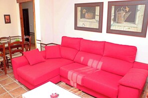 A red sectional sofa in a living room with a tiled floor, a wooden dining table with chairs, and two framed pictures on the wall.